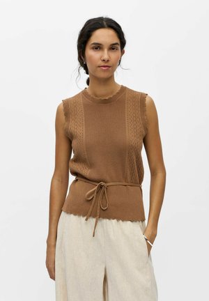 Femme aux cheveux foncés porte un haut sans manches en tricot marron avec ceinture nouée à la taille et un pantalon ample beige clair, debout sur fond blanc.