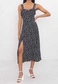 Černé květinové šaty s bílým květinovým vzorem, rýškovaným výstřihem a širokými ramínky; mají boční rozparek a splývavý A-silhouette.