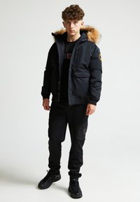 Giubbotto a collo alto invernale nero con cappuccio in pelliccia sintetica, chiusura con zip e bottoni, due tasche frontali; abbinato a pantaloni neri e sneaker nere.