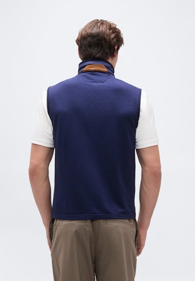 Άνδρας που φοράει λευκό πουκάμισο με κοντά μανίκια, σκούρο μπλε αμάνικο γιλέκο με καφέ λεπτομέρεια στο γιακά και μπεζ παντελόνι, όρθιος με την πλάτη προς τα εμπρός σε λευκό φόντο.