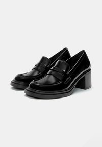 Ein Paar schwarze Lackleder-Blockabsatzslipper mit abgerundetem Zehenbereich und Riemendetail, auf einem schlichten weißen Hintergrund präsentiert.