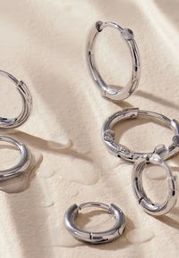 Silberne Creolen in verschiedenen Größen, mit einer glatten, glänzenden Oberfläche und Scharnierverschlüssen. Einige sind teilweise in Wassertropfen getaucht.