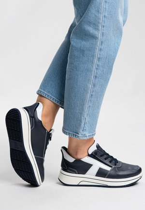 Person wearing light blue jeans and black sneakers with white accents, showing one shoe's sole and the other foot flat on the ground.