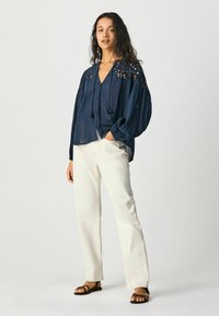 Blusa leggera di colore blu navy con maniche a sbuffo, tagli decorativi sulle spalle, con frange sul collo, abbinata a pantaloni bianchi e sandali.