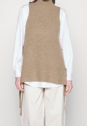 Personne porte un gilet sans manches beige surdimensionné en tricot par-dessus une chemise blanche à manches longues et un pantalon blanc ample sur un fond uni.