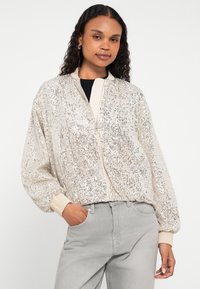 Casaco bomber de cetim em creme com lantejoulas, punhos canelados e fecho de correr frontal. Apresenta um corte descontraído e detalhe de textura na superfície.