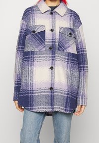 Person, der eine lila und weiße, großkariertete Knopfjacke mit zwei vorderen Klapptaschen trägt, kombiniert mit hellblauen Jeans.