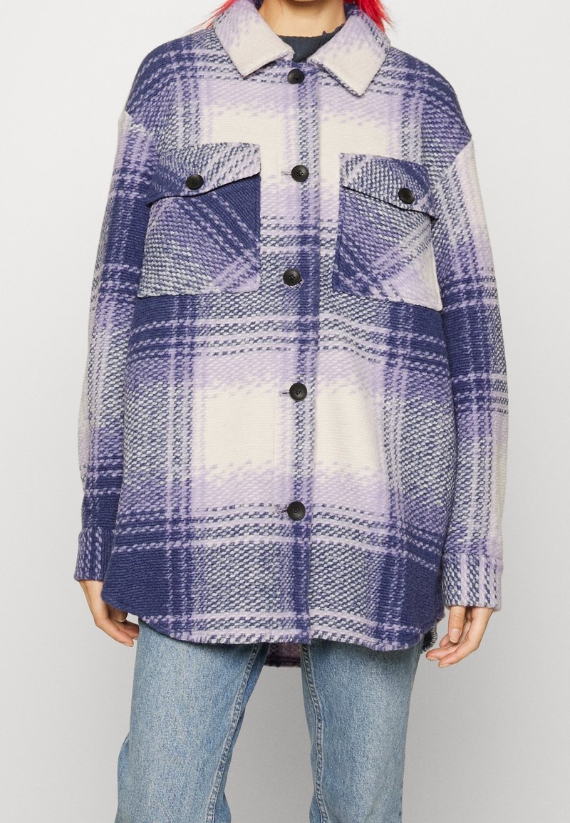Person, der eine lila und weiße, großkariertete Knopfjacke mit zwei vorderen Klapptaschen trägt, kombiniert mit hellblauen Jeans.