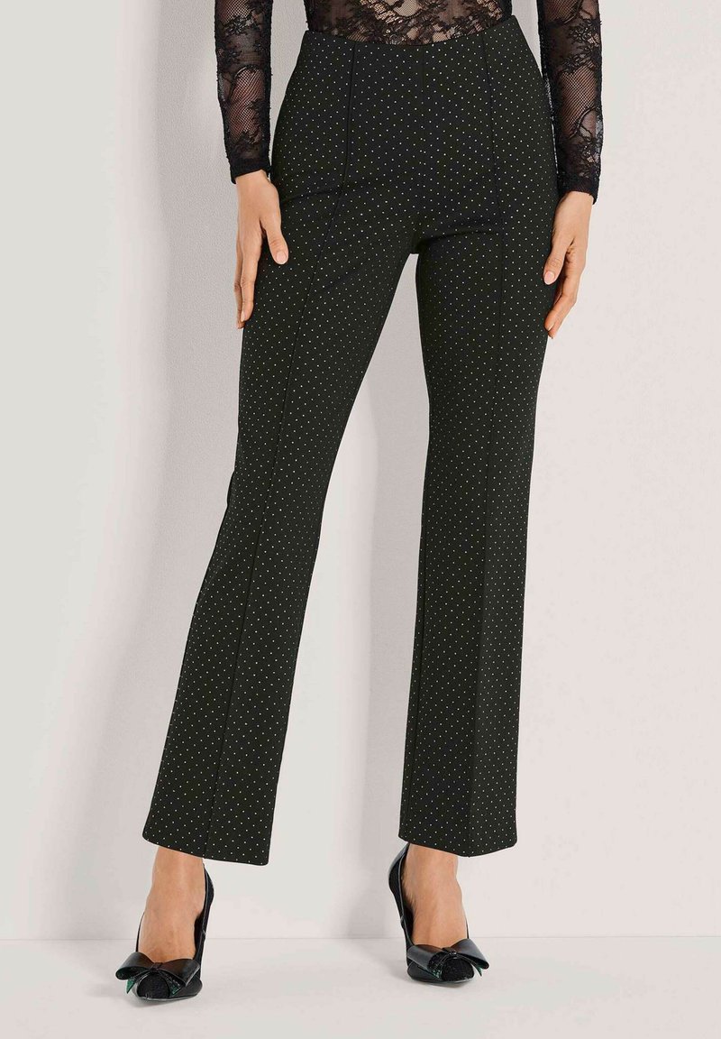 Vrouw in zwarte high-waist broek met kleine witte stippen, gecombineerd met een zwart kanten top met lange mouwen en zwarte strikhakken.