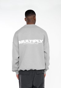 Felpa grigia con spalle scese, polsini e vita a coste. Presenta un grande testo bianco sul retro: "MULTIPLY APPAREL CLUB."