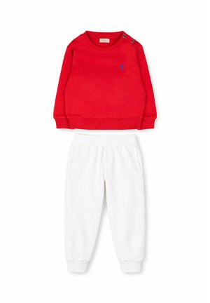 Czerwona bluza dla malucha z długim rękawem, z guzikami na ramieniu i małym niebieskim logo, w połączeniu z białymi dresowymi spodniami ze ściągaczami.