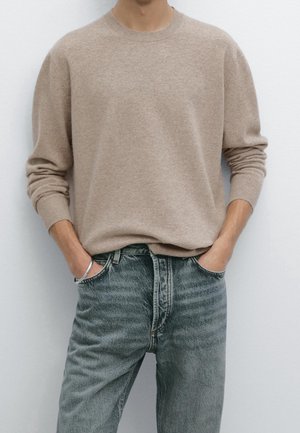 Man die een beige trui met lange mouwen draagt, ingestopt in een hooggesneden, verweekte blauwe spijkerbroek, met handen in de zakken, tegen een witte achtergrond.