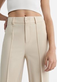 Beżowe, dopasowane spodnie z wysokim stanem, z zapięciem na guziki z przodu, płaskimi zakładkami na przodzie i gładką fakturą.