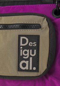 Olivovo zelená obdĺžniková látková taška s textúrovanou čiernou logom nášivkou, tenkým zipsovým vreckom a fialovými akcentmi na vrchnom okraji.