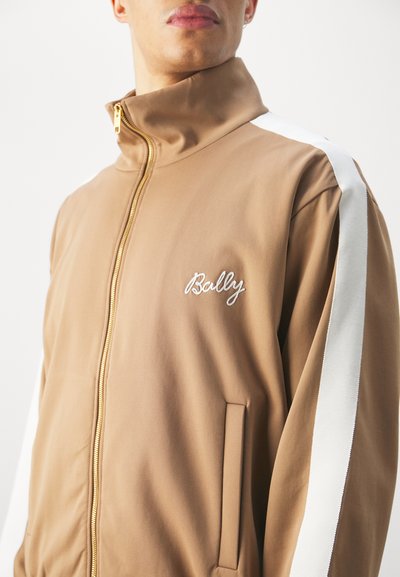 Giacca marrone con zip e colletto alto, strisce laterali bianche, zip dorata e scritta 'Bally' ricamata in bianco sul lato sinistro del petto. Texture liscia.