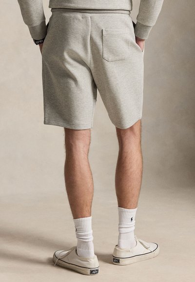 Homme portant un short gris clair, des chaussettes blanches et des baskets écrues, debout les mains dans les poches sur un fond uni.