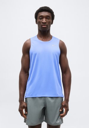 Mężczyzna stojący przodem, ubrany w jasnoniebieską sportową koszulkę bez rękawów i szare spodenki na jednolitym białym tle.