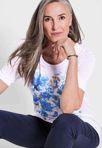 Witte T-shirt met blauwe bloemenprint; ronde halslijn; korte mouwen. Donkerblauwe broek; gladde textuur; zittende pose, hand op de kin.