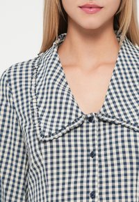 Μπλούζα από ναυτικό και κρεμ καρό με μεγάλο γιακά, μπροστινό κουμπί και ρυτίδες κατά μήκος των άκρων του γιακα. Ελαφριά υφή υφάσματος.