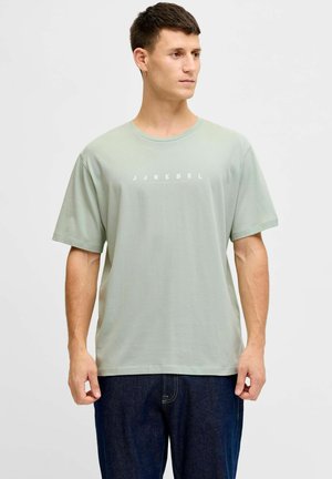 Joven con camiseta verde clara J.JREBEL y vaqueros azul oscuro, mirando hacia un lado sobre un fondo liso.