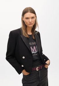 Czarna marynarka dwurzędowa z teksturowanymi klapami, z srebrnymi guzikami, noszona na czarnej koszulce z grafiką i ciemnych dżinsach, zestawiona z fioletowym paskiem.