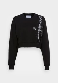 Černá zkrácená mikina s dlouhými rukávy, žebrovanými manžetami a bílým textem "Calvin Klein Jeans" a lesklým logem "CK" na pravé přední straně.