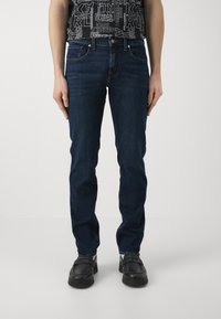 Jeans de color azul oscuro con un diseño de pierna recta, estilo de cinco bolsillos y detalles de costura sutiles. Combinados con zapatos negros.