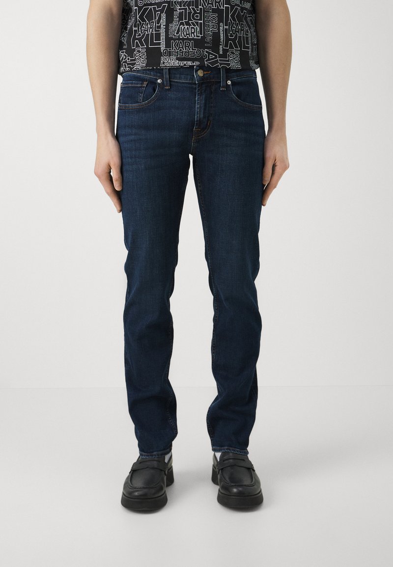 Jeans de color azul oscuro con un diseño de pierna recta, estilo de cinco bolsillos y detalles de costura sutiles. Combinados con zapatos negros.