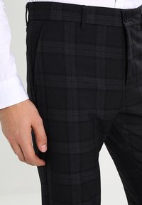 Pantaloni a scacchi neri e grigi con una vestibilità sartoriale, realizzati in un tessuto texture. Presentano una parte anteriore piatta, passanti per la cintura e tasche.