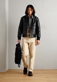 Black leather bomber jacket with front pockets, worn over a light sweater, paired with beige pants and black shoes, holding a black backpack.