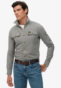 Graues Hemd mit hohem Kragen, zwei Brusttaschen, braunen Knöpfen und einem Aufnäher; kombiniert mit blauen Jeans und einem braunen Gürtel.