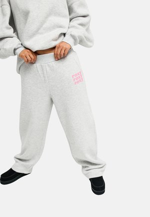Personne tenant la taille d’un pantalon de survêtement gris clair avec texte rose « roxy », associé à un sweat-shirt oversize assorti et des chaussures noires sans lacets.