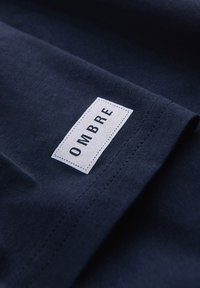 Tissu bleu marine avec une étiquette rectangulaire blanche cousue indiquant « OMBRE » en lettres capitales noires près d'une couture.