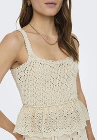 Top sin mangas de color crema hecho de delicado encaje con un diseño de peplum. Presenta bordes ondulados y un patrón floral en la tela.