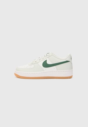 Baskets basses blanches avec le logo Nike vert et semelle couleur gomme, vues de profil sur un fond gris clair uni.