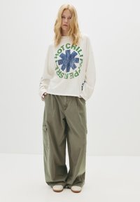 Blonde persoon in een oversized witte lange mouwen shirt met de tekst "Red Hot Chili Peppers" en groene cargobroek, staande voor een effen achtergrond.