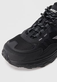 Ténis desportivo preto com sobreposições em malha e sintéticas, colarinho acolchoado, sola texturizada e atacadores refletores contrastantes para visibilidade.