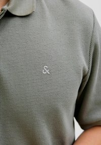 Harmaa tekstuuripolo pusero, jossa on pieni brodeerattu ampersand-logo rinnassa. Lyhyet hihat, pehmeä kangas, klassinen kaulustyyli.