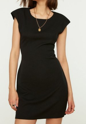Zwart nauwsluitend jurkje met vleermuismouwen, ronde halslijn en een gladde textuur. Voorzien van een gouden ketting met een rechthoekige hanger.