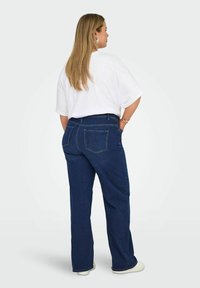 Tumši zili šauri džinsi ar plašām kājām un brīvu piemērījumu, ar šuvju akcentiem un aizmugures kabatām, apvienoti ar baltu pārspīlētu T-kreklu.