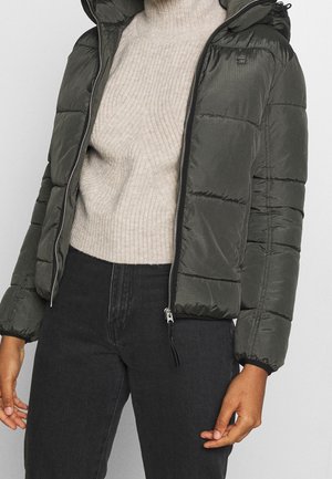 Person mit dunkelgrüner gesteppter Jacke über einem beige gerippten Pullover und schwarzer High-Waist-Jeans, stehend vor einfarbigem Hintergrund.