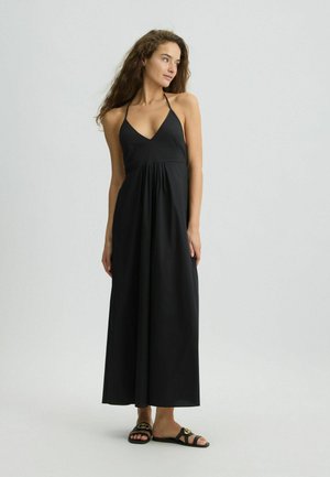 Black maxi dress with a V-neckline, halter straps, and gathered waist. Worn with black slide sandals featuring a gold logo detail.