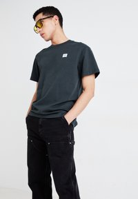 Dark green cotton t-shirt with short sleeves and a small white logo patch, paired with black pants featuring visible stitching and pockets.