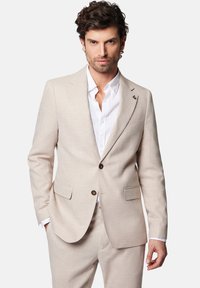 Beżowy lniany garnitur o teksturowanym wykończeniu, jednorzędowa marynarka, dwie przednie kieszenie oraz biała koszula pod spodem. Widoczne detale w postaci brązowych guzików.