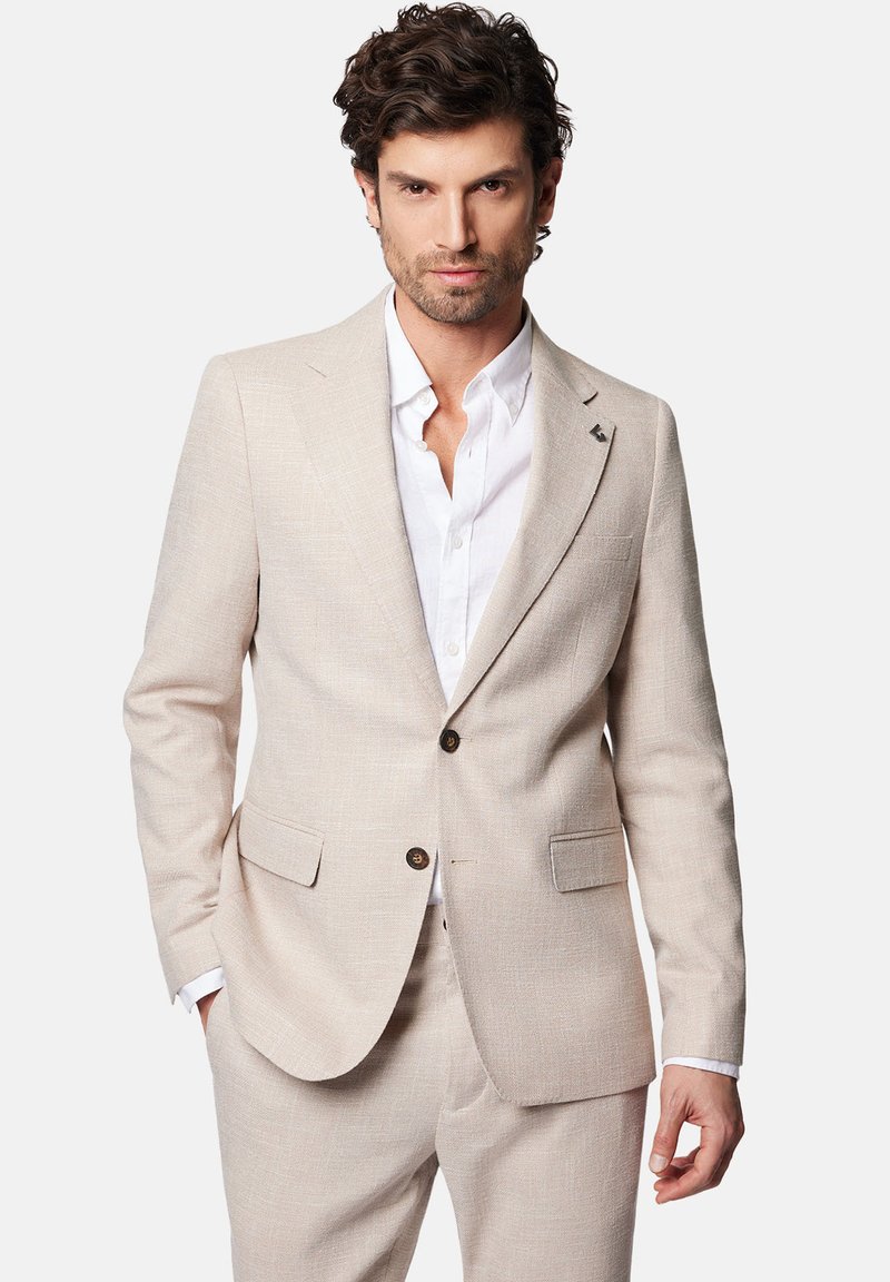 Beżowy lniany garnitur o teksturowanym wykończeniu, jednorzędowa marynarka, dwie przednie kieszenie oraz biała koszula pod spodem. Widoczne detale w postaci brązowych guzików.