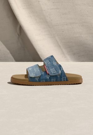 Șlapi cu talpă bej și două curele ajustabile din denim albastru, având un model subtil și o etichetă roșie cu brandul pe lateral.