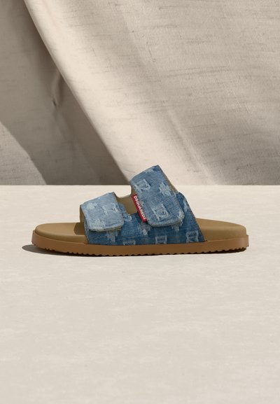 Natikač s masleno podplatom in dvema nastavljivima modrima trakovoma iz džinsa z nežnim vzorcem ter rdečo oznako blagovne znamke na strani.