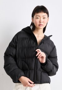 Melna pūkaina jaka ar kapuci un rāvējslēdzēja aizdari, ar kvadrātveida šuvēm un elastīgām aprocēm. Viegls audums ar matētu apdari.