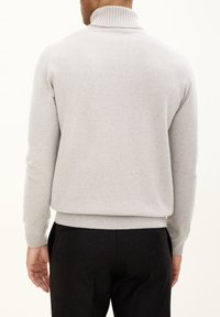 Grauer, gestrickter Pullover mit hohem Rippenkragen und enganliegenden Bündchen, der eine weiche Textur zeigt. Kombiniert mit schwarzen Hosen.