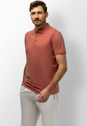 Man draagt een zalmkleurig poloshirt en een witte broek, staand met één hand in zijn zak tegen een egale achtergrond.
